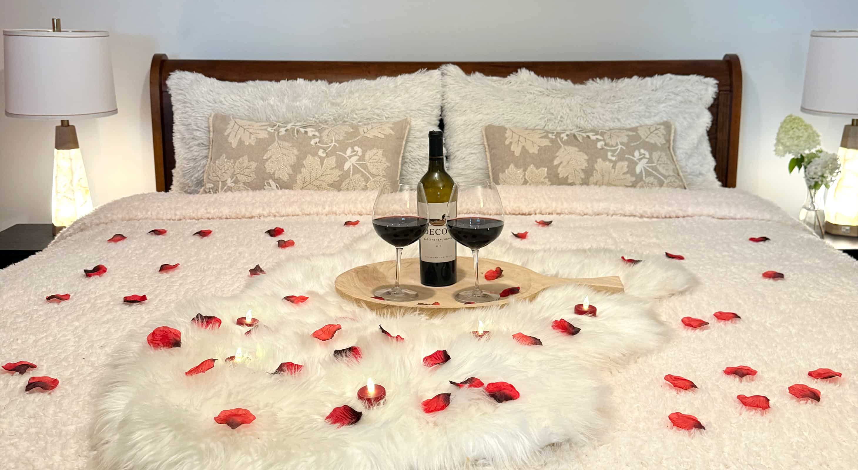 Red wine with wine glasses, rose pedals, and tea lights on a bed