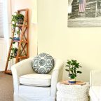 A seating area with two white chairs, a bookshelf, and a basket of blankets in the Riverside studio at Warren Lodge in VT