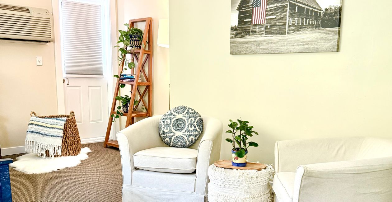 A seating area with two white chairs, a bookshelf, and a basket of blankets in the Riverside studio at Warren Lodge in VT